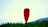 A single red kayak paddle stands upright against a backdrop of a clear light blue sky. Below the paddle, a line of green trees and a distant blue hill create a serene and natural setting.