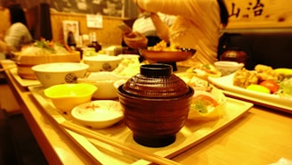 A warmly lit dining setting with various Japanese dishes arranged on wooden trays. In the foreground, a brown wooden bowl is prominently placed, surrounded by small bowls containing different types of food such as salad, rice, and fried items. In the background, people are engaged in conversation.
