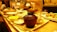 A warmly lit dining setting with various Japanese dishes arranged on wooden trays. In the foreground, a brown wooden bowl is prominently placed, surrounded by small bowls containing different types of food such as salad, rice, and fried items. In the background, people are engaged in conversation.