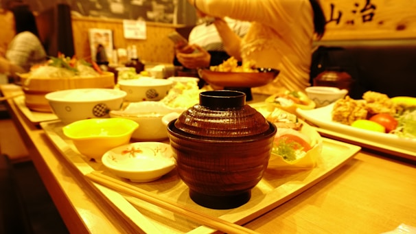 A warmly lit dining setting with various Japanese dishes arranged on wooden trays. In the foreground, a brown wooden bowl is prominently placed, surrounded by small bowls containing different types of food such as salad, rice, and fried items. In the background, people are engaged in conversation.