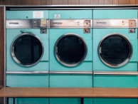 Three teal-colored industrial washing machines with silver accents are lined up side by side against a wooden wall. Each machine features a large circular window and coin slots above, with signs indicating pricing and instructions. Below the machines is a simple wooden bench.
