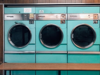 Three teal-colored industrial washing machines with silver accents are lined up side by side against a wooden wall. Each machine features a large circular window and coin slots above, with signs indicating pricing and instructions. Below the machines is a simple wooden bench.