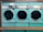 Three teal-colored industrial washing machines with silver accents are lined up side by side against a wooden wall. Each machine features a large circular window and coin slots above, with signs indicating pricing and instructions. Below the machines is a simple wooden bench.