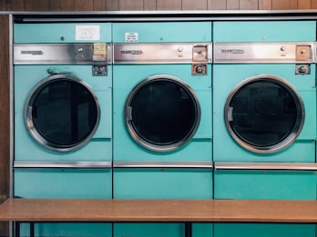 Three teal-colored industrial washing machines with silver accents are lined up side by side against a wooden wall. Each machine features a large circular window and coin slots above, with signs indicating pricing and instructions. Below the machines is a simple wooden bench.