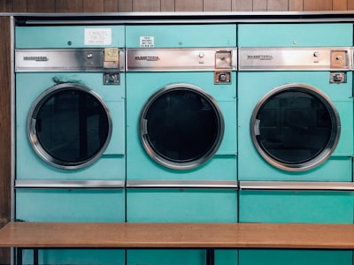Three teal-colored industrial washing machines with silver accents are lined up side by side against a wooden wall. Each machine features a large circular window and coin slots above, with signs indicating pricing and instructions. Below the machines is a simple wooden bench.