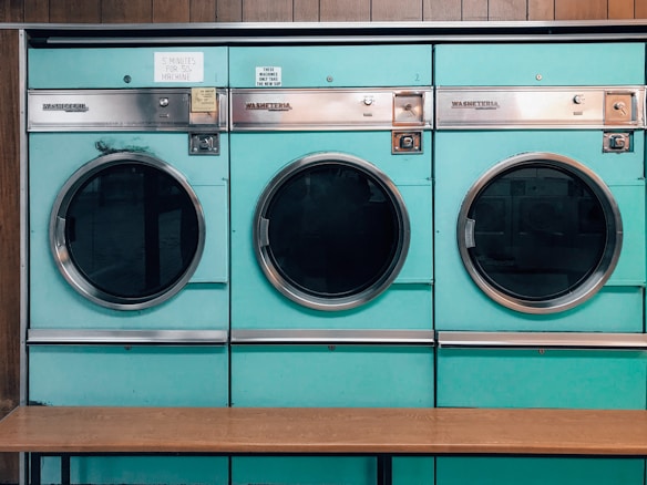 Three teal-colored industrial washing machines with silver accents are lined up side by side against a wooden wall. Each machine features a large circular window and coin slots above, with signs indicating pricing and instructions. Below the machines is a simple wooden bench.