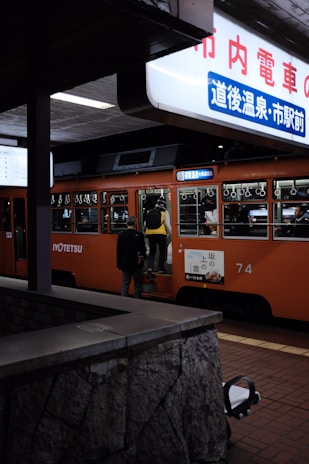 A person boards an orange train with various passengers visible through the windows. A brightly lit sign with Japanese characters hangs above, and there is a stone barrier in the foreground.
