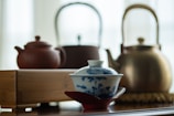 A beautifully crafted porcelain tea cup with blue floral patterns resting on a red saucer in the foreground. In the background, there are three different teapots: one made of clay, one metallic with a bamboo handle, and another in a darker shade. A wooden tea tray is also visible.