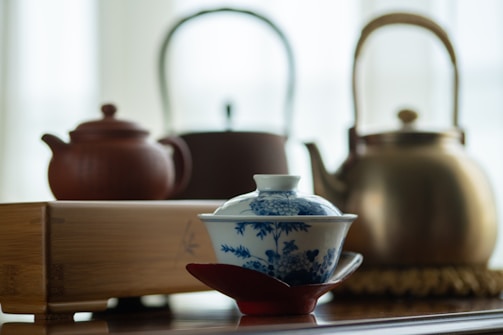 A beautifully crafted porcelain tea cup with blue floral patterns resting on a red saucer in the foreground. In the background, there are three different teapots: one made of clay, one metallic with a bamboo handle, and another in a darker shade. A wooden tea tray is also visible.
