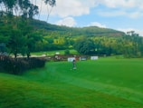 A lush, green golf course set against a backdrop of rolling hills and a clear blue sky. A person is in mid-swing, preparing to hit a golf ball on the manicured grass. Surrounding the area are vibrant trees and some signage, indicating a golf event or tournament.