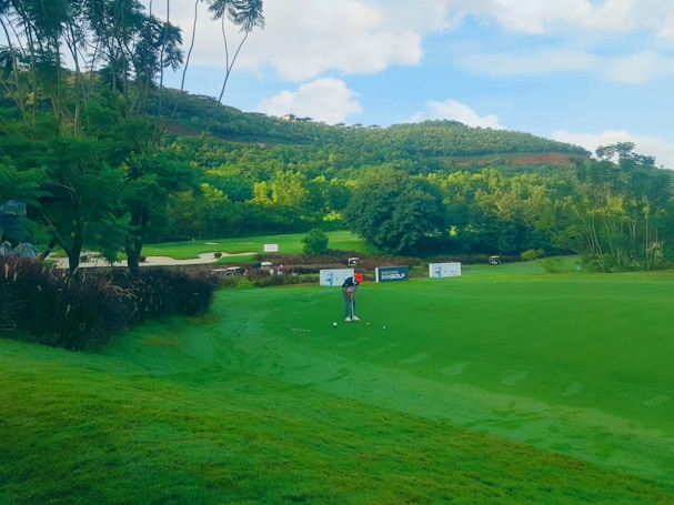 A lush, green golf course set against a backdrop of rolling hills and a clear blue sky. A person is in mid-swing, preparing to hit a golf ball on the manicured grass. Surrounding the area are vibrant trees and some signage, indicating a golf event or tournament.