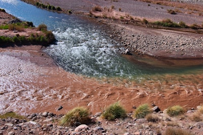 A river with clear, bluish water converges with muddy, brown water, creating a distinctive contrast. The surrounding area features rocky and arid terrain with sparse vegetation, including patches of grass and small shrubs.