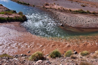 A river with clear, bluish water converges with muddy, brown water, creating a distinctive contrast. The surrounding area features rocky and arid terrain with sparse vegetation, including patches of grass and small shrubs.