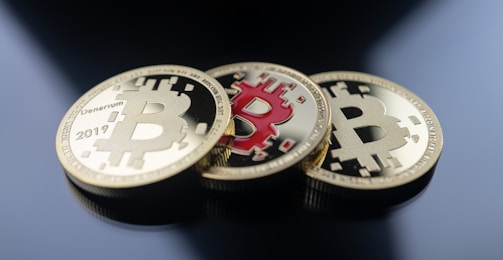 Three physical coins resembling Bitcoin are placed on a reflective dark surface. The coins have a gold color with a large 'B' symbol at the center and circuit-like patterns. One coin stands out due to its red interior.