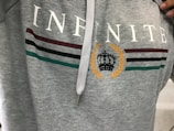 A gray hoodie with the word 'INFINITE' printed in white across the chest. Below the text are three horizontal stripes in maroon, black, and green. A laurel wreath encircles a stylized black crown in the center.