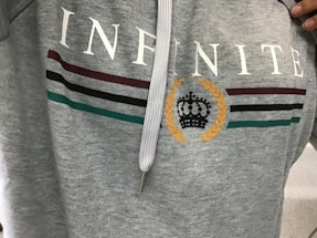 A gray hoodie with the word 'INFINITE' printed in white across the chest. Below the text are three horizontal stripes in maroon, black, and green. A laurel wreath encircles a stylized black crown in the center.
