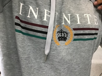 A gray hoodie with the word 'INFINITE' printed in white across the chest. Below the text are three horizontal stripes in maroon, black, and green. A laurel wreath encircles a stylized black crown in the center.