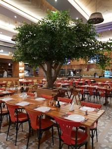 A spacious restaurant interior featuring wooden tables and chairs, with bright red chairs providing a vivid contrast. A large decorative tree stands prominently in the center, adding a touch of nature to the ambiance. Tables are neatly set with plates, cutlery, and napkins, ready for diners. The ceiling has elegant wooden paneling with modern light fixtures adding warmth to the setting. Shelves and a counter are visible in the background, indicating a well-organized dining area.