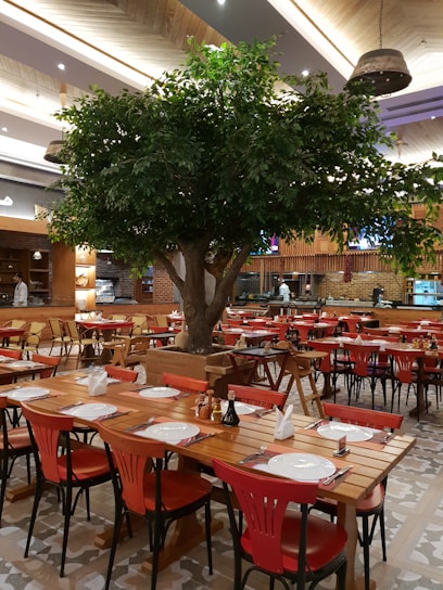 A spacious restaurant interior featuring wooden tables and chairs, with bright red chairs providing a vivid contrast. A large decorative tree stands prominently in the center, adding a touch of nature to the ambiance. Tables are neatly set with plates, cutlery, and napkins, ready for diners. The ceiling has elegant wooden paneling with modern light fixtures adding warmth to the setting. Shelves and a counter are visible in the background, indicating a well-organized dining area.