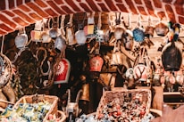 A collection of various decorative bells and trinkets is displayed, hanging from a rustic wooden framework. Each bell is uniquely designed, with intricate patterns and vibrant colors. The setting has a warm, rustic charm with wicker baskets filled with colorful ornaments below, set against a backdrop of red brick and wooden elements.