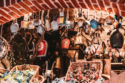 A collection of various decorative bells and trinkets is displayed, hanging from a rustic wooden framework. Each bell is uniquely designed, with intricate patterns and vibrant colors. The setting has a warm, rustic charm with wicker baskets filled with colorful ornaments below, set against a backdrop of red brick and wooden elements.