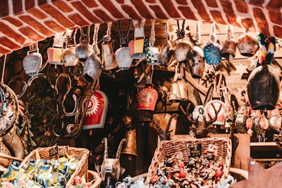 A collection of various decorative bells and trinkets is displayed, hanging from a rustic wooden framework. Each bell is uniquely designed, with intricate patterns and vibrant colors. The setting has a warm, rustic charm with wicker baskets filled with colorful ornaments below, set against a backdrop of red brick and wooden elements.