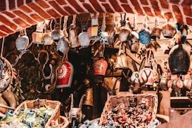 A collection of various decorative bells and trinkets is displayed, hanging from a rustic wooden framework. Each bell is uniquely designed, with intricate patterns and vibrant colors. The setting has a warm, rustic charm with wicker baskets filled with colorful ornaments below, set against a backdrop of red brick and wooden elements.