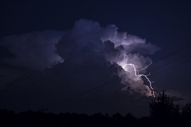 A powerful lightning bolt illuminates a large, dark cloud formation against a night sky. Electrical lines cut across the image, adding a sense of scale and context to the storm. The scene is dramatic, with a stark contrast between the bright lightning and the dark surroundings.