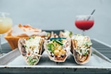 A trio of tacos filled with various ingredients such as shredded lettuce, cheese, jalapeños, pineapple, and meat, arranged on a metal tray. In the background, there is a basket of tortillas or chips and two drinks, one yellowish and the other reddish with straws.