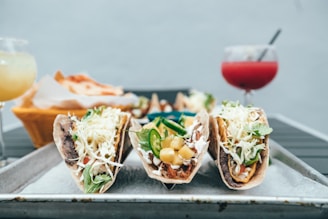 A trio of tacos filled with various ingredients such as shredded lettuce, cheese, jalapeños, pineapple, and meat, arranged on a metal tray. In the background, there is a basket of tortillas or chips and two drinks, one yellowish and the other reddish with straws.