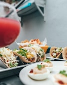 A close-up view of a variety of tacos filled with ingredients such as lettuce, cheese, and pineapple, arranged in metal taco holders. In the background, there are crispy tortilla chips and a red drink in a glass, with deviled eggs garnished with herbs on a plate in the foreground.