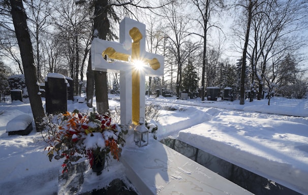 A snowy cemetery scene with a large cross prominently displayed in the foreground. Sunlight streams through the cross, creating a glowing effect. A bouquet of colorful flowers is placed next to the cross, adding vibrancy to the wintry environment. Bare trees and gravestones are visible in the background.