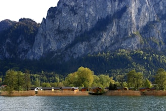 Small houses are nestled among lush trees at the base of a towering rocky mountain. The scene is bordered by a tranquil lake in the foreground, reflecting the natural beauty and diversity of the landscape.