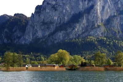 Small houses are nestled among lush trees at the base of a towering rocky mountain. The scene is bordered by a tranquil lake in the foreground, reflecting the natural beauty and diversity of the landscape.
