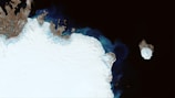 A satellite image shows a large expanse of icy terrain, characterized by predominantly white and brown patches along the edges, with dark blue and black waters surrounding it. An isolated, rounded icy landmass appears in the water to the right, slightly separated from the main ice field.
