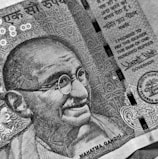 A detailed black and white close-up of a currency note featuring an illustration of a bespectacled man with a smile. The note includes intricate patterns and text in both English and another language. A promise to pay in the sum of one hundred rupees is prominently displayed alongside official signatures and seals.