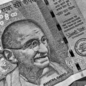 A detailed black and white close-up of a currency note featuring an illustration of a bespectacled man with a smile. The note includes intricate patterns and text in both English and another language. A promise to pay in the sum of one hundred rupees is prominently displayed alongside official signatures and seals.