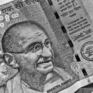 A detailed black and white close-up of a currency note featuring an illustration of a bespectacled man with a smile. The note includes intricate patterns and text in both English and another language. A promise to pay in the sum of one hundred rupees is prominently displayed alongside official signatures and seals.
