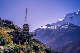 A rugged mountain landscape with a prominent snow-capped peak under a clear blue sky. In the foreground, a rocky outcrop is adorned with a small stack of stones and a prayer flag, set against lush green vegetation and colorful wildflowers.