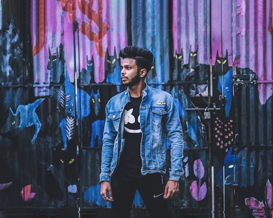 A person wearing a denim jacket and black t-shirt stands in front of a colorful graffiti wall depicting cat-like figures with glowing eyes. The wall is painted with vibrant shades of pink, blue, and purple, creating an urban artistic backdrop.