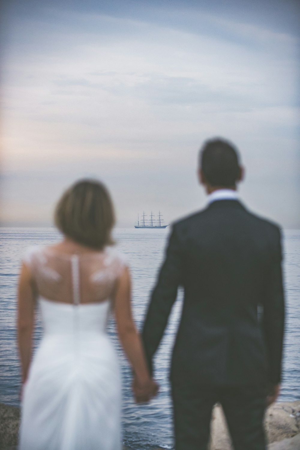 woman wearing whtie wedding gown and man black wearing black suit jacket and pants looking at ocean