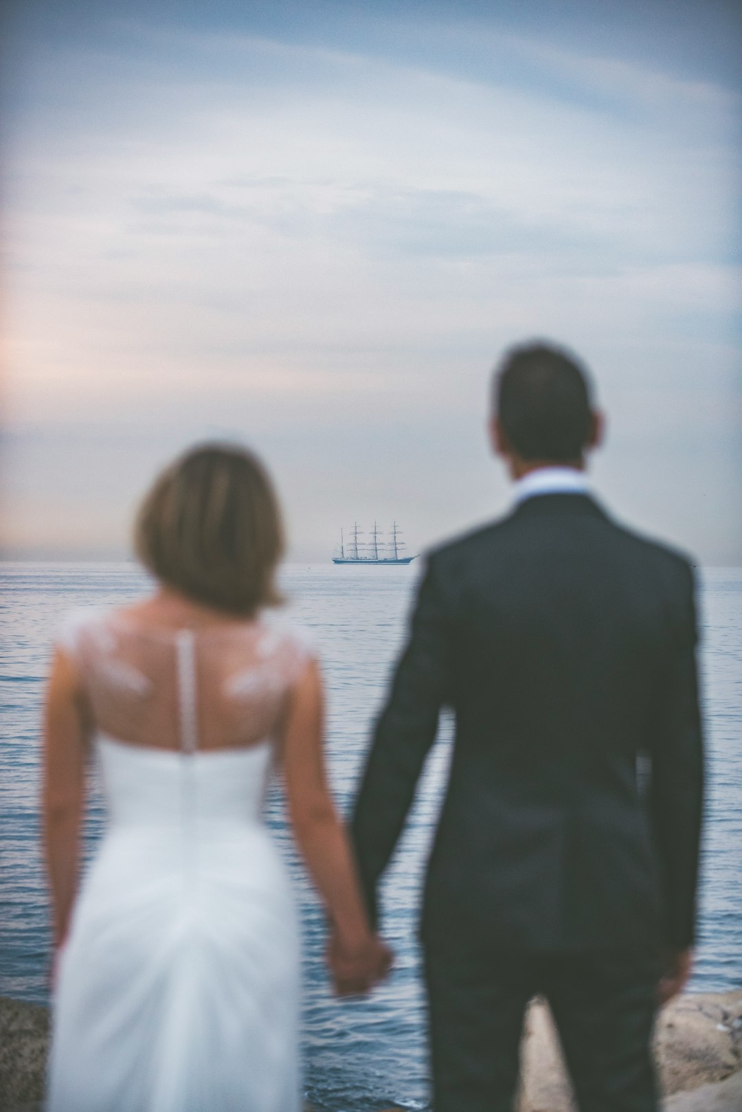 woman wearing whtie wedding gown and man black wearing black suit jacket and pants looking at ocean