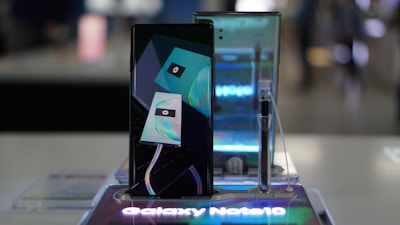 A smartphone is displayed on a stand, showcasing its sleek design. Behind it, a reflective surface enhances its modern look. The device's screen features a stylized graphic, and a stylus is placed to the side.