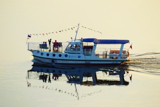 A small boat with a canopy is navigating through calm waters. It has several people on board, including a man standing near the edge. The boat is adorned with small triangular flags and features a distinct reflection in the water. The surrounding water is tranquil, allowing the reflection to be clearly visible.