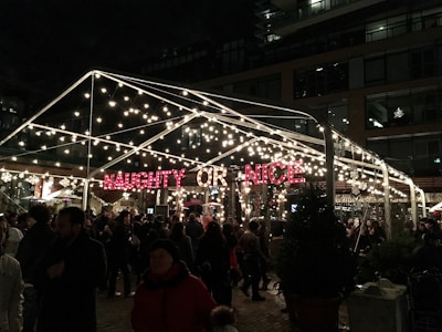 A festive outdoor setting with a transparent tent adorned with numerous twinkling lights and a bright red sign displaying the words 'NAUGHTY OR NICE'. Dense crowds of people are gathered underneath, enjoying the evening ambiance. The background features a multi-story building with illuminated windows, enhancing the lively atmosphere.