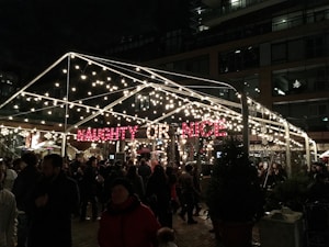 A festive outdoor setting with a transparent tent adorned with numerous twinkling lights and a bright red sign displaying the words 'NAUGHTY OR NICE'. Dense crowds of people are gathered underneath, enjoying the evening ambiance. The background features a multi-story building with illuminated windows, enhancing the lively atmosphere.