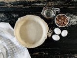 A pie crust is placed in a pie pan on a rustic dark wooden surface, surrounded by three eggs, a bottle of vanilla extract, a cup of pecans, and a measuring cup filled with water. A white kitchen towel with black stripes is also visible.