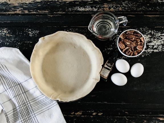 A pie crust is placed in a pie pan on a rustic dark wooden surface, surrounded by three eggs, a bottle of vanilla extract, a cup of pecans, and a measuring cup filled with water. A white kitchen towel with black stripes is also visible.