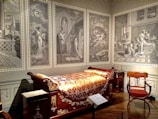 A room furnished with an opulent wooden bed adorned with gold patterns and an intricate blanket. The walls are decorated with large, monochromatic murals depicting classical scenes with figures in ancient attire. An elegant wooden chair with a red cushion is positioned near the bed, and various classical elements and decorations fill the room.