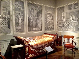 A room furnished with an opulent wooden bed adorned with gold patterns and an intricate blanket. The walls are decorated with large, monochromatic murals depicting classical scenes with figures in ancient attire. An elegant wooden chair with a red cushion is positioned near the bed, and various classical elements and decorations fill the room.